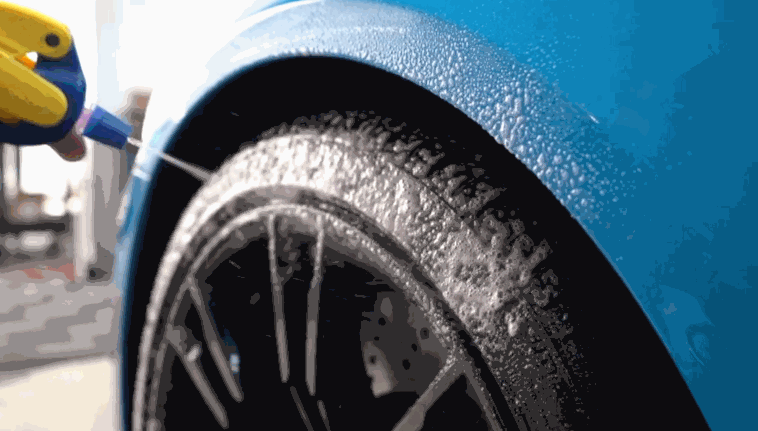 A vibrant blue sports car’s tire gets coated in cleaning foam as a detailer sprays it using a yellow and blue bottle, preparing the wheel for a deep clean.