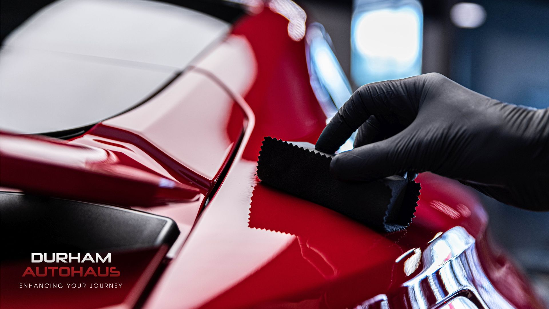A gloved hand applies a ceramic coating to the glossy red surface of a car using a black cloth applicator. The high-shine finish reflects the shop lights, with “Durham Autohaus – Enhancing Your Journey” branding in the corner.