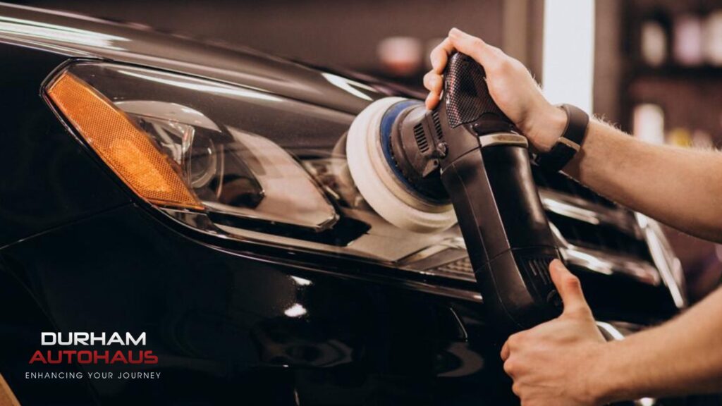 Close-up of a car headlight being polished with a power buffer by a technician, showcasing the detailing process at Durham Autohaus. The black vehicle’s surface reflects the surroundings, and the shop’s branding is visible in the corner.
