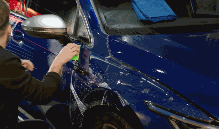 Technician uses a green squeegee to apply PPF over the driver-side fender and mirror of a blue car.